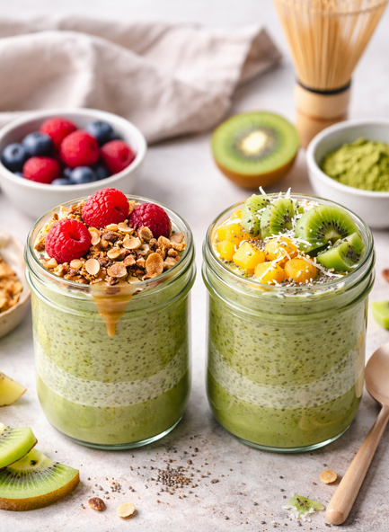 Zwei Gläser cremiger Matcha Chia Pudding mit frischen Beeren, Kiwi, Mango, Granola und Kokos als Topping auf hellem Hintergrund angerichtet.