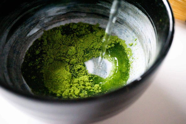Dunkle Tasse mit Matcha-Tee, der mit Wasser übergossen wird.