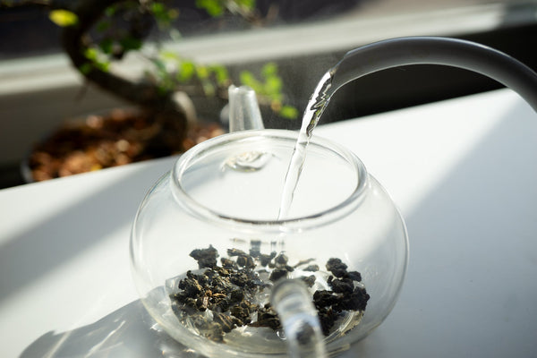 Eine Glas-Teekanne mit Teeblättern, in die gerade Wasser gegossen wird