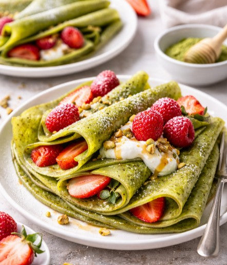Frisch gebackene Matcha Crêpes auf einem Teller, gefüllt mit Erdbeeren und Joghurt, garniert mit Pistazien, Puderzucker und einem Schuss Honig.