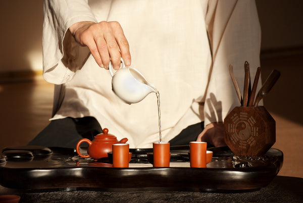 Ein schwarzes Tablett mit drei terracottafarbenen Teetassen, in denen Tee aufgegossen wird